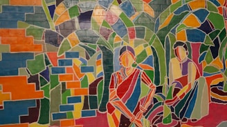 A vibrant mural depicts two abstract, stylized figures sitting amidst lush, leafy greenery. The artwork is composed of geometric shapes filled with a spectrum of colors, resembling a stained glass design. The figures appear to be wearing traditional attire.