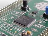 A close-up view of a green printed circuit board featuring various electronic components, including an integrated circuit chip marked with identification codes, resistors, and connectors. The board is intricately designed, showing numerous paths and soldered points.