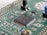 A close-up view of a green printed circuit board featuring various electronic components, including an integrated circuit chip marked with identification codes, resistors, and connectors. The board is intricately designed, showing numerous paths and soldered points.
