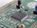 A close-up view of a green printed circuit board featuring various electronic components, including an integrated circuit chip marked with identification codes, resistors, and connectors. The board is intricately designed, showing numerous paths and soldered points.