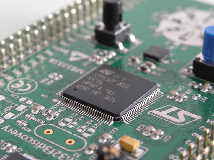 A close-up view of a green printed circuit board featuring various electronic components, including an integrated circuit chip marked with identification codes, resistors, and connectors. The board is intricately designed, showing numerous paths and soldered points.