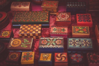A collection of intricately painted wooden boxes with colorful floral designs is displayed on a surface. Each box varies in size and shape, showcasing unique patterns in vibrant colors such as red, orange, green, and blue. The boxes feature detailed artwork, combining geometric shapes and elaborate decorations that reflect a traditional or folk art style.