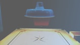 A carrom board is positioned beneath a ceiling lamp, which casts a soft light over the scene. The board is marked with standard carrom designs, including corner pockets and a central circle. The surface of the board appears smooth and ready for a game.