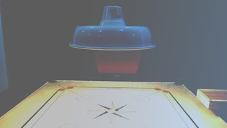 A carrom board is positioned beneath a ceiling lamp, which casts a soft light over the scene. The board is marked with standard carrom designs, including corner pockets and a central circle. The surface of the board appears smooth and ready for a game.