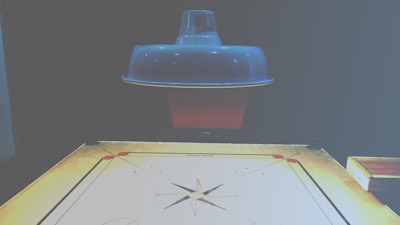 A carrom board is positioned beneath a ceiling lamp, which casts a soft light over the scene. The board is marked with standard carrom designs, including corner pockets and a central circle. The surface of the board appears smooth and ready for a game.