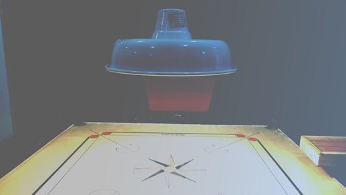 A carrom board is positioned beneath a ceiling lamp, which casts a soft light over the scene. The board is marked with standard carrom designs, including corner pockets and a central circle. The surface of the board appears smooth and ready for a game.