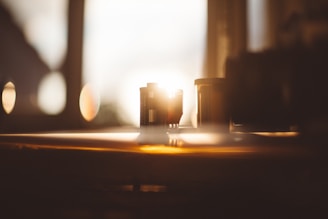 A soft focus captures a sunlit scene with vintage film canisters resting on a wooden surface. Warm light streams through a window, creating an inviting and nostalgic ambience.