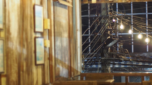 A wooden interior with framed pictures on the wall is seen in the foreground. In the background, there is a view of a complex network of electrical cables and power lines illuminated by several light bulbs. The setting seems to blend elements of rustic decor and urban infrastructure.