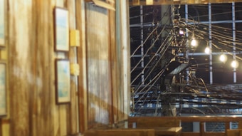 A wooden interior with framed pictures on the wall is seen in the foreground. In the background, there is a view of a complex network of electrical cables and power lines illuminated by several light bulbs. The setting seems to blend elements of rustic decor and urban infrastructure.