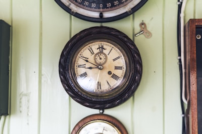 A vintage wall clock with Roman numerals encased in a decorative dark wooden frame is mounted on a light green paneled wall. Beside it hangs a small metal winding key. Another clock or barometer is partially visible beneath it.