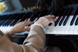 An elderly person's hands are playing a piano. The person is wearing a pearl bracelet. The piano keys are black and white, and there is a logo visible on the piano.