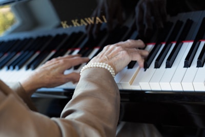 An elderly person's hands are playing a piano. The person is wearing a pearl bracelet. The piano keys are black and white, and there is a logo visible on the piano.