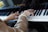 An elderly person's hands are playing a piano. The person is wearing a pearl bracelet. The piano keys are black and white, and there is a logo visible on the piano.