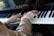An elderly person's hands are playing a piano. The person is wearing a pearl bracelet. The piano keys are black and white, and there is a logo visible on the piano.
