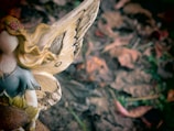 A close-up view of a decorative fairy figurine with intricate details. The figurine has golden hair adorned with flowers, wearing a blue and yellow dress. It features large, ornate wings with a brown texture. The background consists of blurred brown leaves, suggesting an outdoor, earthy setting.