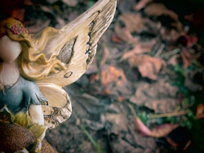 A close-up view of a decorative fairy figurine with intricate details. The figurine has golden hair adorned with flowers, wearing a blue and yellow dress. It features large, ornate wings with a brown texture. The background consists of blurred brown leaves, suggesting an outdoor, earthy setting.