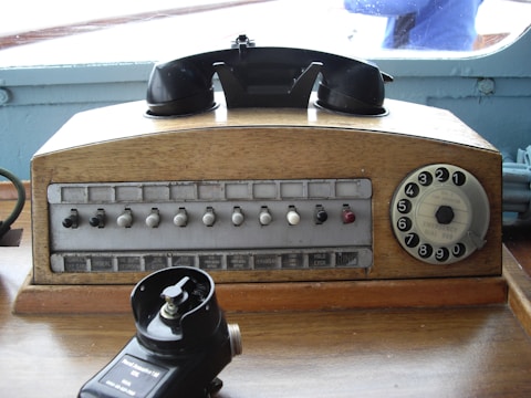 A vintage rotary dial telephone with a wooden casing, featuring a black handset resting on top. The front panel shows several labeled buttons and switches, possibly for different lines or functions, along with a rotary dial on the right side. In front, there is a black device, likely an old hand microphone, placed on a wooden surface.