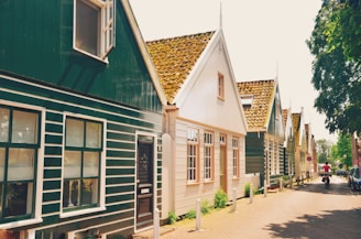 A row of traditional European-style houses with steeply pitched roofs line a narrow brick-paved street. The buildings feature wooden exteriors, painted in shades of green and beige, with white trim around the windows and doors. The roofs are covered with brown terracotta tiles. There are some trees providing shade on the right side of the street, and a person riding a bicycle can be seen further down the street.