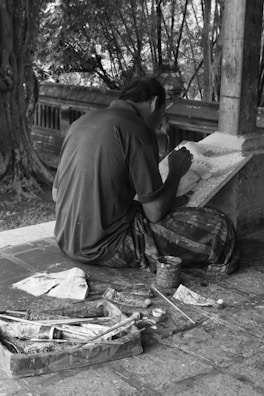 A person is seated on the ground, deeply focused on painting a detailed artwork on a canvas or board. The setting appears to be outdoors, surrounded by trees and rustic elements. Art supplies, such as brushes and a tray with paint tubes, are scattered on the stone floor beside the artist.