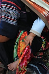A child's hand and foot are partially visible under a colorful, patterned fabric. The fabric is draped over an adult's arm and leg, which are clad in garments with intricate patterns and vivid colors. The child's skin is light against the dark and colorful textiles.
