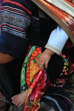 A child's hand and foot are partially visible under a colorful, patterned fabric. The fabric is draped over an adult's arm and leg, which are clad in garments with intricate patterns and vivid colors. The child's skin is light against the dark and colorful textiles.