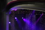 A stage setup with bright purple spotlights illuminating a space. The background consists of numerous small, twinkling lights, resembling stars in a dimly lit environment. The ceiling has layered arches adding depth and texture to the scene.