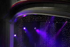 A stage setup with bright purple spotlights illuminating a space. The background consists of numerous small, twinkling lights, resembling stars in a dimly lit environment. The ceiling has layered arches adding depth and texture to the scene.