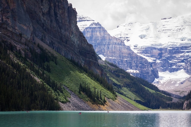 A serene lake surrounded by steep rocky mountains covered with patches of snow and lush green forests. The water is a calm turquoise color, contrasting with the rugged, towering cliffs. A few kayakers can be seen on the lake, enjoying the tranquil scenery.