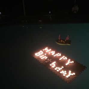 Floating on still water, numerous small lights form the phrase 'Happy Birthday'. Adjacent to the lights is a small decoration shaped like a ship with tiny American flags, adding a celebratory feel.
