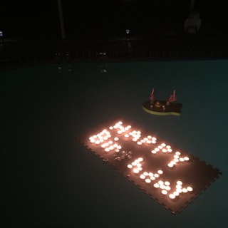 Floating on still water, numerous small lights form the phrase 'Happy Birthday'. Adjacent to the lights is a small decoration shaped like a ship with tiny American flags, adding a celebratory feel.