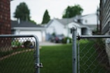 A chain-link fence gate partially open, leading to a grassy backyard area. Several houses with gray roofs and white walls are visible in the background, partially blurred. Trees and bushes are present, adding greenery to the suburban neighborhood setting.