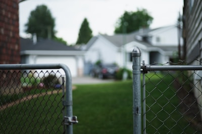 A chain-link fence gate partially open, leading to a grassy backyard area. Several houses with gray roofs and white walls are visible in the background, partially blurred. Trees and bushes are present, adding greenery to the suburban neighborhood setting.