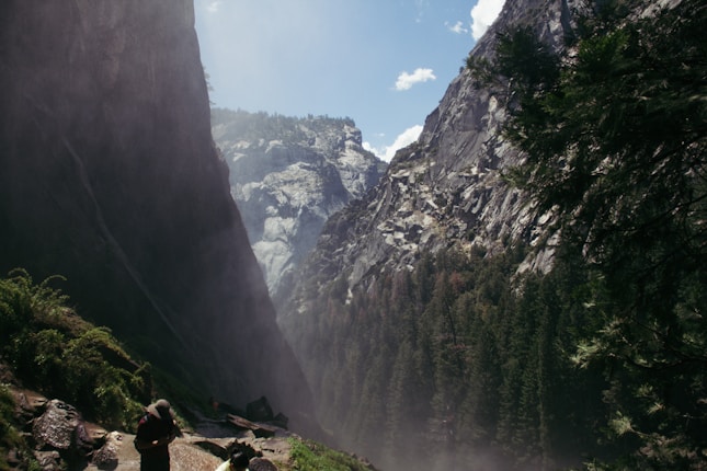 A dramatic mountain scene with steep rocky cliffs and a misty atmosphere. Tall evergreen trees line the valley, while a hiker walks along a misty trail at the bottom left, creating a sense of scale and adventure.