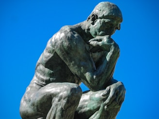 A bronze sculpture of a muscular man in a contemplative pose, resting his chin on one hand with his body leaned forward. The sky is clear and bright blue in the background.