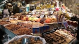 A bustling market stall displays an array of goods, including nuts, dried fruits, chocolates, and candies. Various types of bottles, possibly containing oils or vinegars, are stacked in the background. The scene is busy with customers browsing, while a vendor in a white apron assists them. The display is colorful and densely packed, showcasing a rich variety of offerings.