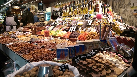 A bustling market stall displays an array of goods, including nuts, dried fruits, chocolates, and candies. Various types of bottles, possibly containing oils or vinegars, are stacked in the background. The scene is busy with customers browsing, while a vendor in a white apron assists them. The display is colorful and densely packed, showcasing a rich variety of offerings.