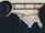 A set of vintage clothing items hangs on a wooden coat rack against a patterned, pale wallpaper. The clothing includes a black jacket with a large decorative hat and a black dress with an intricate white lace collar. The wooden hangers have floral designs or printed text.