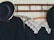A set of vintage clothing items hangs on a wooden coat rack against a patterned, pale wallpaper. The clothing includes a black jacket with a large decorative hat and a black dress with an intricate white lace collar. The wooden hangers have floral designs or printed text.