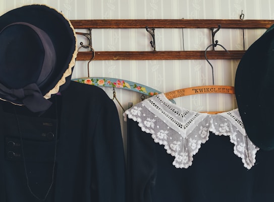 A set of vintage clothing items hangs on a wooden coat rack against a patterned, pale wallpaper. The clothing includes a black jacket with a large decorative hat and a black dress with an intricate white lace collar. The wooden hangers have floral designs or printed text.