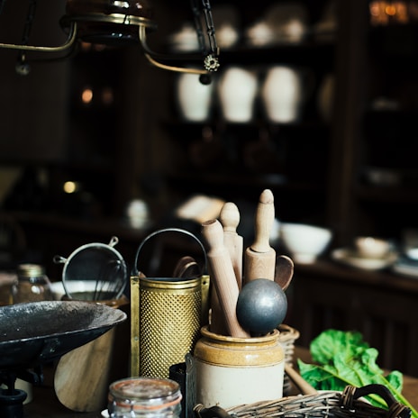 A rustic kitchen scene featuring a collection of kitchen utensils including wooden rolling pins, a metal grater, a sieve, and a mortar on a wooden counter. In the background, there are shelves with pottery and bowls, and a bunch of fresh greens is placed in the corner. The lighting is dim, creating an antique and cozy ambiance.
