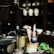 A rustic kitchen scene featuring a collection of kitchen utensils including wooden rolling pins, a metal grater, a sieve, and a mortar on a wooden counter. In the background, there are shelves with pottery and bowls, and a bunch of fresh greens is placed in the corner. The lighting is dim, creating an antique and cozy ambiance.