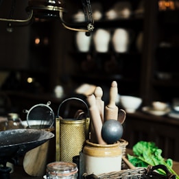 A rustic kitchen scene featuring a collection of kitchen utensils including wooden rolling pins, a metal grater, a sieve, and a mortar on a wooden counter. In the background, there are shelves with pottery and bowls, and a bunch of fresh greens is placed in the corner. The lighting is dim, creating an antique and cozy ambiance.