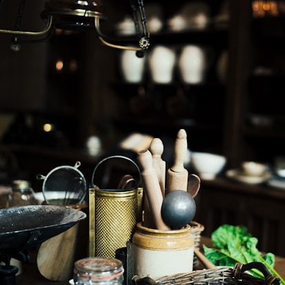 A rustic kitchen scene featuring a collection of kitchen utensils including wooden rolling pins, a metal grater, a sieve, and a mortar on a wooden counter. In the background, there are shelves with pottery and bowls, and a bunch of fresh greens is placed in the corner. The lighting is dim, creating an antique and cozy ambiance.