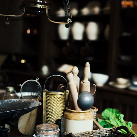 A rustic kitchen scene featuring a collection of kitchen utensils including wooden rolling pins, a metal grater, a sieve, and a mortar on a wooden counter. In the background, there are shelves with pottery and bowls, and a bunch of fresh greens is placed in the corner. The lighting is dim, creating an antique and cozy ambiance.