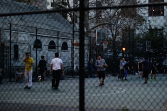 A group of people playing basketball on an outdoor court surrounded by a tall wire fence. The court is located in an urban setting, with buildings and trees visible in the background. Some players are actively engaged in the game, while others stand around the perimeter. The atmosphere seems relaxed and informal.