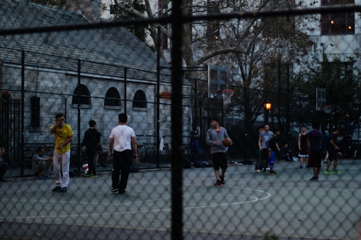 A group of people playing basketball on an outdoor court surrounded by a tall wire fence. The court is located in an urban setting, with buildings and trees visible in the background. Some players are actively engaged in the game, while others stand around the perimeter. The atmosphere seems relaxed and informal.
