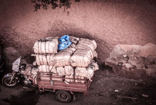 A motorized cart is heavily loaded with large bales wrapped in beige fabric, secured with black cords. There's a single blue bale on top, and the cart is parked against a weathered, textured wall with peeling paint.