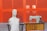 A white torso mannequin is seated on a wooden chair next to an old-fashioned sewing machine placed on a wooden table. The background consists of bright orange panels, giving a bold contrast to the scene. A pair of headphones rests beside the sewing machine.