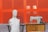 A white torso mannequin is seated on a wooden chair next to an old-fashioned sewing machine placed on a wooden table. The background consists of bright orange panels, giving a bold contrast to the scene. A pair of headphones rests beside the sewing machine.