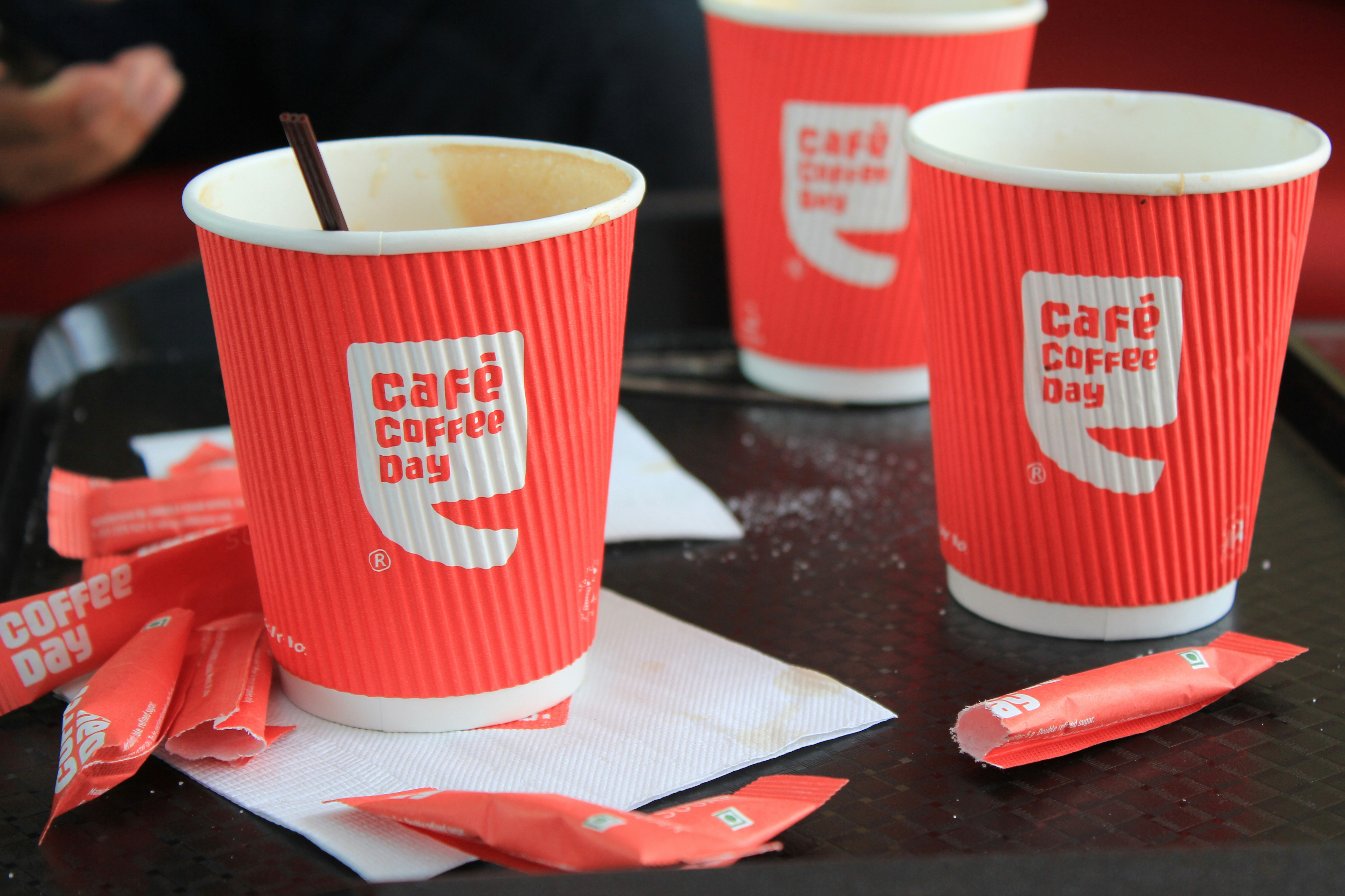 Three red paper cups with the 'Café Coffee Day' logo are placed on a tray. There are also several empty sugar sachets scattered around on white napkins.
